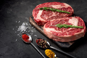 Poster - Two raw rib eye steak with seasonings on the dark stone background prepared for cooking. Marbled beef top view