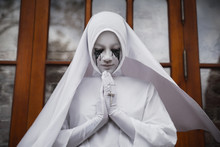 A Gloomy Portrait Of A Ghostly Pale Nun In White Robes With Black Eye Sockets And Black Smudges Under Them, Amid Gloomy Gothic Architecture. Halloween, Horror.