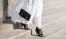 Fashionable Bag Close-up In Female Hands.Girl Walks In The City Outdoors. Stylish Modern And Feminine Image, Style. Girl In A White Light Dress With A Black Bag And Black Boots, Or Heeled Shoes.