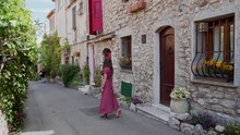 Beautiful Young Girl In Red Polka-dot Dress Standing Near Red Door Of Ancient Stone House In Romantic French City Antibes. Distant Plan