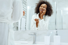 Skin Care. Woman Applying Face Cream Looking In Bathroom Mirror