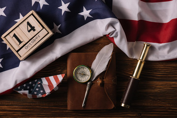 Sticker - top view of wooden calendar with October 14 date, paper boat, nib, compass, telescope and leather notebook on wooden surface with American national flag