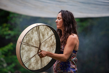 Wall Mural - Diverse people enjoy spiritual gathering A free-spirited and confident mystical lady is seen wearing colorful clothes as she plays a sacred native drum during a sacred ritual in natural surroundings.