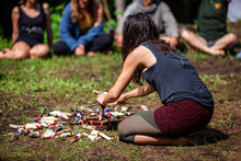 Diverse People Enjoy Spiritual Gathering A Bohemian Style Woman Is Seen Crafting Sacred Objects During A Woodland Retreat Celebrating Shamanism And Native Cultures With Blurry People In Background.