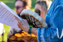 Sacred Drums During Spiritual Singing. A Close Up View On The Hands Of A Spiritual Guide, Holding A Music Book And Native Drum During A Cultural Music Celebration In A Park During Summer.
