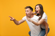 Shocked young couple in white t-shirts posing isolated on yellow orange background. People lifestyle concept. Mock up copy space. Giving piggyback ride to joyful sitting on back pointing finger aside.