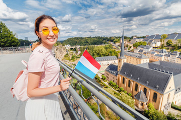 Wall Mural - A happy Asian traveller girl holds the flag of Luxembourg and admires the Grund area from the observation deck. Tourism, recreation and life in the country.