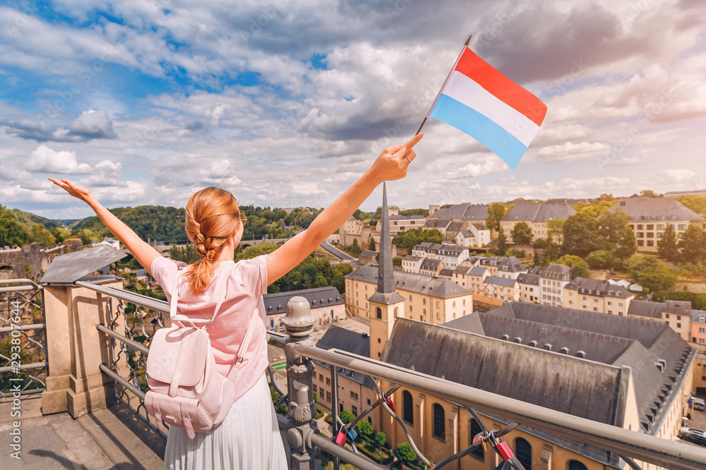 Obraz na płótnie A happy traveller girl holds the flag of Luxembourg and admires the Grund area from the observation deck. Tourism, recreation and life in the country. w salonie