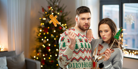 Wall Mural - christmas, photo booth and holidays concept - gloomy couple in ugly sweaters posing with party props over home room background