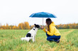 A woman with her dog sitting under a large umbrella in the rain. A young girl crouched in front of her dog on a green field in the background of the forest, rainy autumn. Outdoors or outside