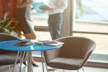 Close up shot of chair and blue coffee table with notebooks lying on it in modern office. Two co-workers looking at documents in the background. Horizontal shot. Selective focus