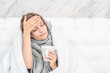 © Ermolaev Alexandr - Sick young woman with warm scarf sitting on the bed under blanket, holds cup and checks the temperature by touching the forehead. Empty space for text