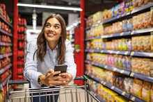 Smiling Woman At Supermarket. Happy Woman At Supermarket. Beautiful Young Woman Shopping In A Grocery Store/supermarket. Shopping Lists In App Format