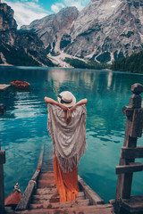 Girl in a straw hat on the background of the turquoise lake with wooden boats in mountains. Safari style blonde woman back view. Dolomites Alps, lago di Braies, Italy