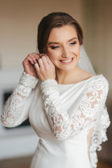 Wall Mural - Close up portrait of beautiful bride standing by the window at home and tying on earrings. Charming bride in white wedding dress