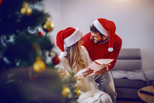 Handsome Caucasian Man Giving Christmas Present To His Loving Girlfriend. Both Are Dressed In Sweaters And Having Santa Hat On Heads. Christmas Holidays.