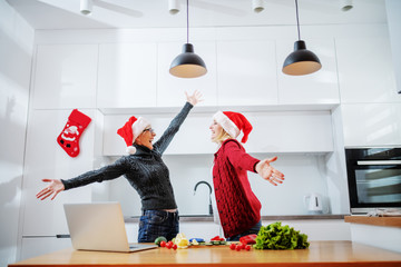 Wall Mural - Overjoyed senior woman standing in kitchen with her pregnant daughter. Both having wide opened arms and santa hats on heads. On kitchen counter are laptop and vegetables.