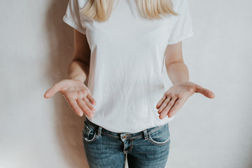 Wall Mural - Blond hair woman posing near a light wall. Beautiful young caucasian girl. Hand gestures. Emotion. Casual clothing. Studio model in work. Strong woman, future is female. Jeans and white blank t-shirt