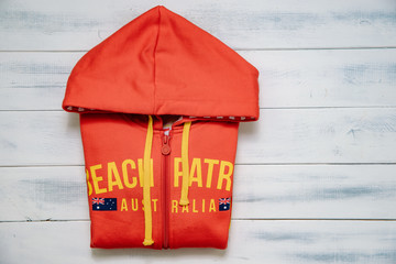 Wall Mural - A red blouse for a lifeguard from a beach in Australia. On the wooden table lies a red sweatshirt and a lifeguard cap. The concept of saving life at the seaside, problems over water.