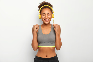 Studio shot of happy African American woman raises clenched fists, celebrates successful won game, dressed in sports bra, listens music via headphones, has slim body curly hair. Fitness female relaxes