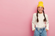 Studio Shot Of Happy Asian Woman With Tender Smile, Keeps Both Hands In Pockets On Jeans, Wears Yellow Hat, White Jumper, Has Two Pigtails Poses Over Rosy Wall Blank Space For Your Advertising Content