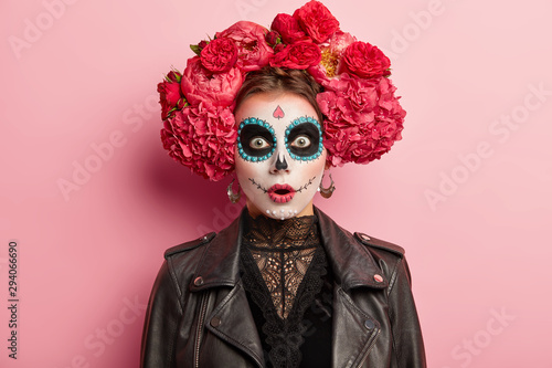Fotomural Shocked terrified young woman has scary ghost face, wears artistic makeup for Day of Dead holiday, wears black leather jacket, models over rosy studio background