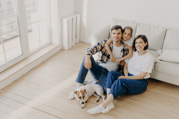 Lovely European family of father, mother and their daughter sit on floor near sofa in spacious white room, pedigree dog lies near, have moving day, change place of living, express happiness.