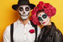 Zombie Portrait. Dead Woman And Man Wears Skull Makeup, Painted For Halloween, Look At Camera Surprisingly, Dressed In Black And White Outfit For All Saint Day, Isolated Over Yellow Background.