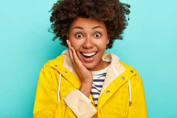 Headshot of good looking African American woman touches cheeks, looks gladfully being in high spirit, dressed in waterproof raincoat, isolated over blue background, gets good news. Season concept