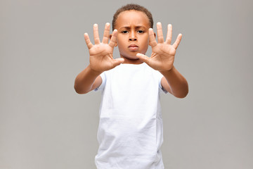 Wall Mural - Portrait of serious Afro American little boy dressed in white t-shirt frowning having grumpy facial expression, showing all ten fingers on both hands at camera, making No gesture or Stop sign