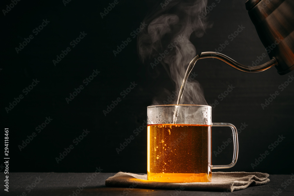 The process of brewing tea, pouring hot water from the kettle into the Cup, steam coming out of the mug, water droplets on the glass, black background - obrazy, fototapety, plakaty 