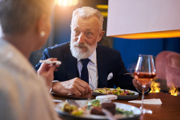 Senior caucasian bearded male sits and look at woman in restaurant in hotel. Elegant speaking man in tuxedo. Blonde woman in white blazer