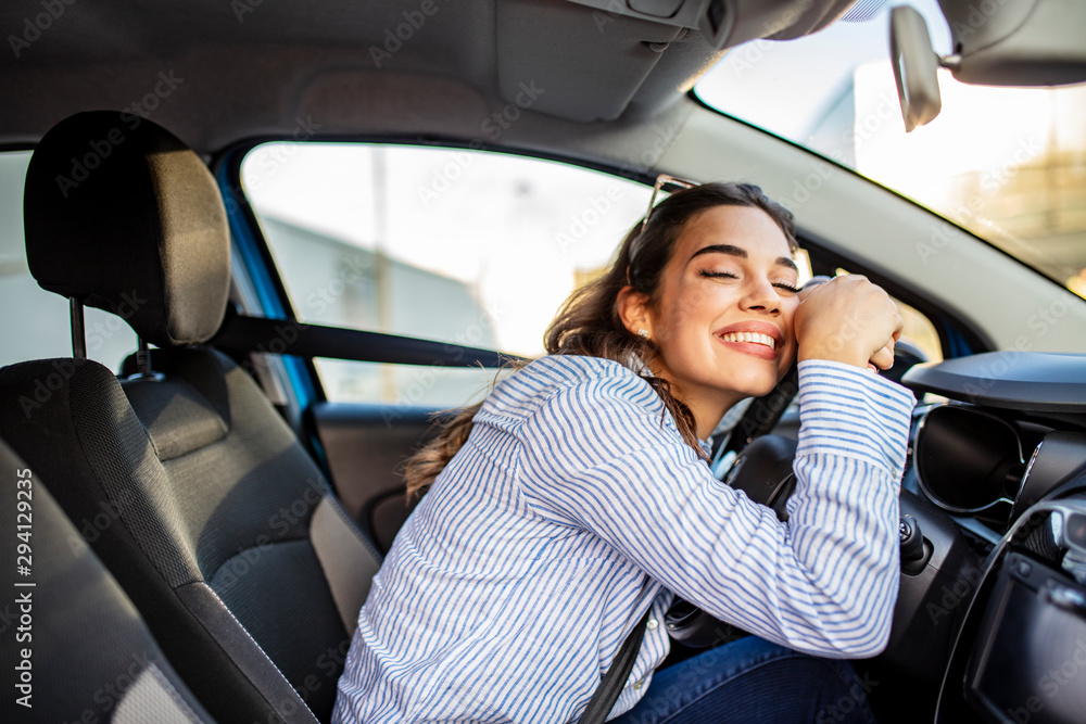 Young Woman Embracing Her New Car. Excited young woman and her new car indoors. Young and cheerful woman enjoying new car hugging steering wheel sitting inside - obrazy, fototapety, plakaty 