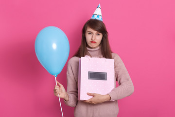 Close up portrait of young beautyful darkhaired woman with gift and blue balloon posing isolated over background. Lady dresses rosy sweater and birthday hat. Celebration and holiday concept.