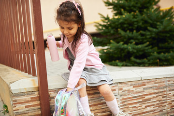 Outdoors image of cute little girl preschooler with pink glass bottle of water and backpack sitting outside. Happy child pupil relaxing outside after preschool lessons. People, education concept