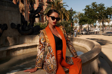 Fashion style outdoor portrait. Elegant woman in colorful orange costume, sunglasses and black shoes sitting on stone fence of fountain on a sunny evening in tropical city