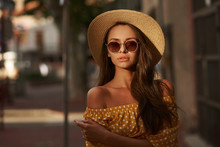 Closeup Outdoor Portrait Of Young Beautiful Caucasian Young Woman With Long Brunette Hair Wearing Yellow Polka Dot Dress, Sunglasses And Thatch Hat. Summer Sunny Evening Street