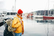 © leonidkos - Back view of carefree man traveller with touristic rucksack wearing yellow coat and rad hat standing on berth and looking toward scandinavian city. Tourist walking the small authentic city among ship
