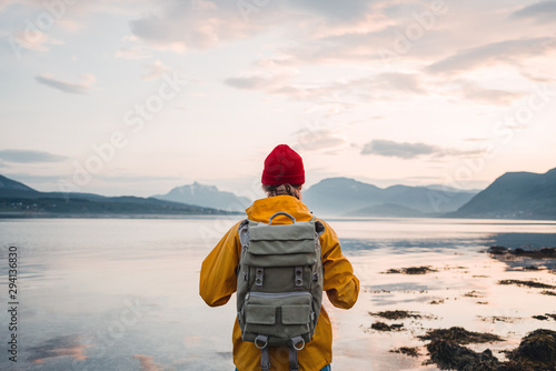 Cuadros en Lienzo Back view of male tourist with rucksack standing on coast in front of great mountain massif while journey