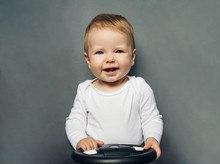 Close-up Portrait Of A Nine-month-old Baby Girl With Blond Hair And Blue Eyes, Dressed In A White Bodysuit On A Gray Background.
