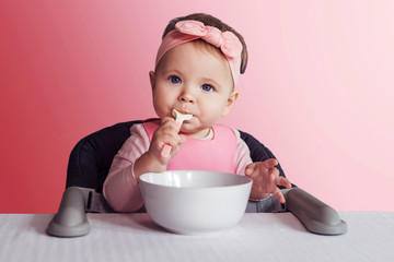 Nine-month-old baby girl in pink bandage sits at white table in highchair, eats herself with spoon from bowl. Pink background.