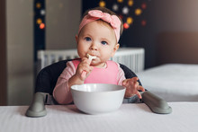 Nine-month-old Smiling Baby Girl Sits At White Table In Highchair And Eats Herself With Spoon From Bowl. Blurred Background.