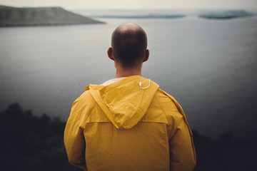 Traveler in yellow raincoat standing on cliff and looking at lake in rainy windy day. Wanderlust and travel concept. Hipster man hiking in Norway on foggy day. Atmospheric moment