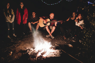 Hipster man playing on acoustic guitar and singing song with friends travelers at big bonfire at night camp in the forest. Group of people chilling at fire in the evening, camping near lake