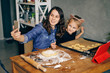 Mother and daughter making video call from smartphone while cooking holiday cookies in winter season at home