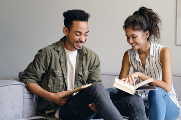 Young smiling African American man and pretty Asian woman joyfully reading book together in co-working space