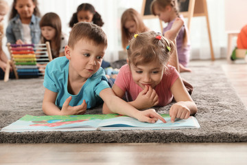 Poster - Cute kids reading book on floor while other children playing together in kindergarten