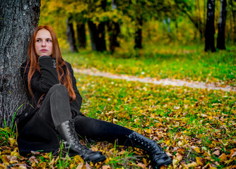 portrait of young teenager redhead girl with long hair sitting on the ground near a tree in black clothes and boots