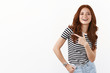© Cookie Studio - Carefree modern young female student in striped t-shirt with ginger curly hairstyle, smiling self-assured, pointing finger left, help find copy space, recommend advertisement, white background