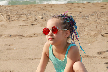 cute girl in fashionable red glasses and colored pigtails sitting on the beach